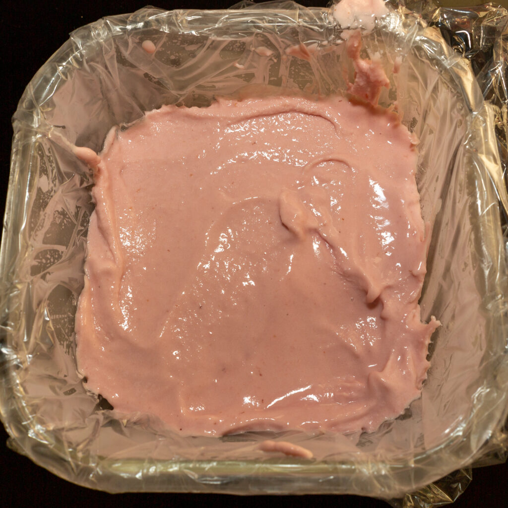Fertiges Himbeer-Eis als unterste Schicht in einer eckigen Form, die mit Frischhaltefolie ausgeschlagen ist.